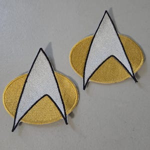 May include: Two embroidered fabric patches featuring a gold oval background with a white arrowhead shape centred on top. Each patch has a distinct black stitched border outlining the arrowhead and the oval shape, creating a sharp, clean design.