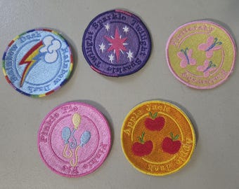 May include: Five circular embroidered patches featuring character icons. One light blue with a rainbow lightning bolt, one purple with a pink star, one yellow with three pink butterflies, one pink with three balloons, and one orange with three red apples.
