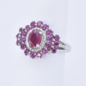 May include: A silver ring with a large oval-shaped ruby surrounded by a halo of smaller purple gemstones.