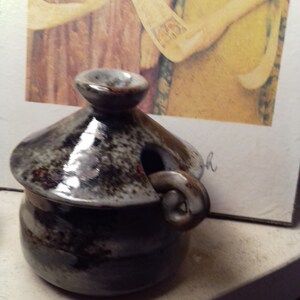 Puede incluir: Un pequeño tarro de cerámica gris y marrón con tapa y asa. El tarro tiene un esmalte moteado y un asa curva única.