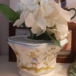 Op de afbeelding: Witte keramische pot met een handgeschilderd ontwerp van een meisje in een blauwe jurk en gele bloemen. De pot heeft een gegolfde rand en is versierd met vlinders.