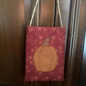 May include: A burgundy fabric wall hanging with a stitched pumpkin applique. The pumpkin is a patchwork of orange and tan fabric. The background has a floral pattern. A natural twine loop is attached for hanging.