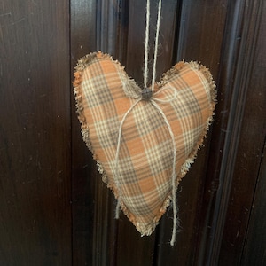 May include: A heart-shaped fabric ornament with a plaid pattern in shades of orange, brown, and cream. The edges are frayed, and it hangs from a twine string. The ornament is tied with twine at the top.