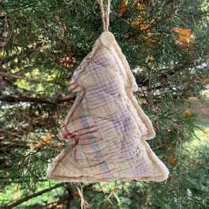 May include: A handmade quilted Christmas tree ornament in beige, purple, and red plaid fabric. The ornament is hanging from a tree branch.