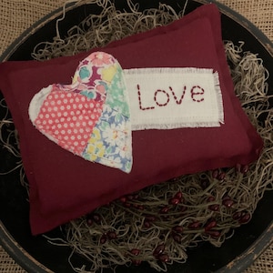 May include: A burgundy pillow with a patchwork heart and the word "Love" embroidered on a white patch. The heart features floral and polka dot patterns in various colors. The pillow is placed in a rustic bowl with decorative elements.