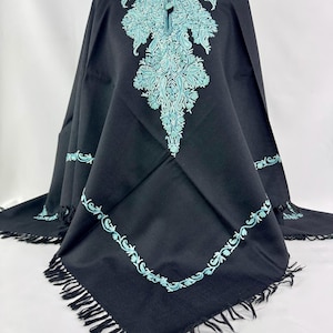 Op de afbeelding: Zwarte cape met ingewikkelde turquoise borduursels rond de halslijn, het midden en de zoom. De cape heeft een vierkante vorm met franjes langs de onderrand. Het borduurwerk heeft een bloemen- en paisley-ontwerp.