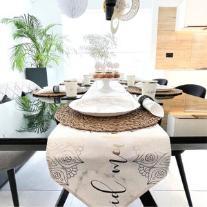 May include: A white table runner with a black and gold design that says "Eid Mubarak". The runner is on a black table with a white plate and a woven placemat. There are white cups and napkins on the table.