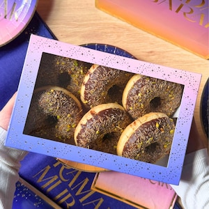Puede incluir: Una caja de seis donuts cubiertos de chocolate con chispas y pistachos. Los donuts están en una caja con ventana, con un diseño degradado azul y rosa. La caja está sobre un plato azul y dorado.