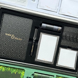Op de afbeelding: Een zwart-gouden briefpapierset met een notitieboek, pen, plaknotities, paperclips en tape. Het notitieboek en de tape hebben een geometrisch patroon. De pen en plaknotities hebben de tekst "Sabr & Shukr".