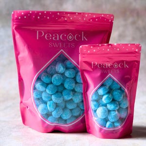 Puede incluir: Dos bolsas rosas de caramelos Peacock Sweets. Las bolsas tienen una ventana transparente en forma de lágrima que muestra caramelos azules. Las bolsas tienen un estampado de lunares blancos en la parte superior y las palabras "Peacock Sweets" en blanco.