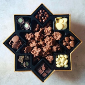 Può includere: Una scatola a forma di stella con bordi dorati, piena di un assortimento di cioccolatini. La scatola è divisa in sezioni contenenti varie caramelle al cioccolato, tra cui tartufi, noci ricoperte e dischi di cioccolato. I cioccolatini sono in tonalità marroni e bianche.