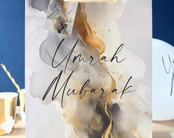 May include: A white gift bag with a gold and gray abstract design. The bag has a white handle and a tag that says "Umrah Mubarak".