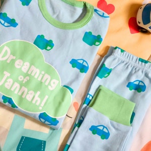 May include: A light blue pajamas set with a green trim. The top has a green cloud with the text "Dreaming of Jannah!" and a pattern of small blue cars. The pants have a similar pattern of blue cars.