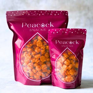 Puede incluir: Dos bolsas de aperitivos de color magenta con el texto "Peacock Snacks" en blanco. Las bolsas tienen una ventana transparente en forma de lágrima que revela aperitivos de color naranja. Las bolsas están selladas y tienen un patrón de puntos en la parte superior.