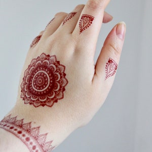 May include: A hand adorned with intricate henna designs in a deep red hue. The designs include a large, detailed mandala on the palm, smaller patterns on the fingers, and a decorative band on the wrist. The skin tone is fair, and the background is a soft gray.