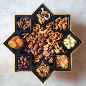 Può includere: Un vassoio nero a forma di stella con bordi dorati, pieno di un assortimento di noci, frutta secca, pretzel e caramelle ricoperte di cioccolato. Il vassoio è diviso in scomparti, ognuno contenente uno spuntino diverso. Una disposizione visivamente accattivante.