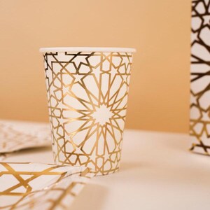 May include: White paper cup with a gold geometric pattern. The cup has a cylindrical shape and a white rim. The gold pattern is a repeating starburst design. The cup is part of a set of party supplies.