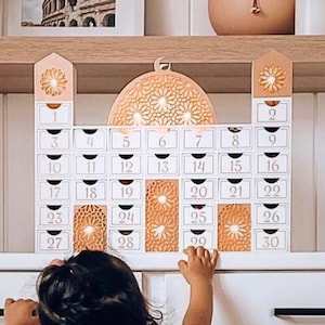 May include: A white wooden advent calendar shaped like a mosque with small drawers numbered 1-30. The top features a decorative dome and two towers with floral cutouts. A child's hands reach for the calendar.