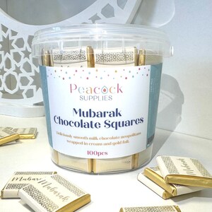 May include: A clear plastic container filled with 100 individually wrapped milk chocolate squares. The squares are wrapped in gold foil and have Arabic calligraphy that reads "Mubarak". The container has a label that reads "Peacock Supplies Mubarak Chocolate Squares Deliciously smooth milk chocolate neopolitans wrapped in cream and gold foil. 100pcs".