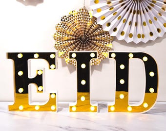 May include: Illuminated gold and black letters spelling "EID" with small white lights. The letters are set against a backdrop of white and gold paper fans. The overall theme is festive and celebratory.