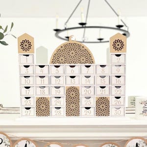 May include: A white wooden advent calendar shaped like a mosque, with 30 numbered drawers. The calendar features decorative cutouts and a domed top. The numbers are in a gold color.