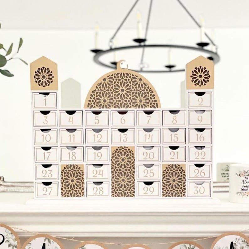 May include: A white wooden advent calendar shaped like a mosque, with 30 numbered drawers. The calendar features decorative cutouts and a domed top. The numbers are in a gold color.
