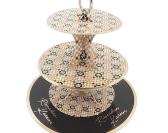 May include: Three-tiered cake stand with a gold and black geometric pattern. The stand has a handle on top and says "Ramadan Kareem" on the bottom tier.