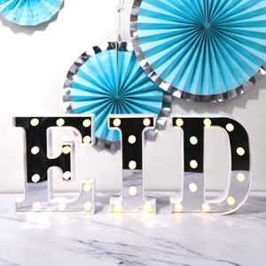 May include: Silver mirrored letters spelling "EID" with white lights. The letters are on a white marble surface. There are three blue and silver paper fans in the background.