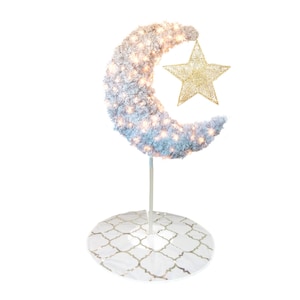 May include: A decorative, illuminated crescent moon and star. The moon is covered in white material and adorned with small lights. A gold star hangs from the moon. The base is white with a gold pattern.