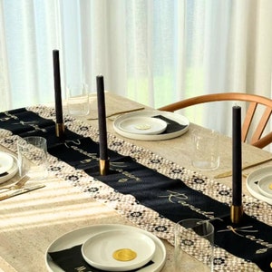 May include: A black and white table runner with gold lettering that says "Ramadan Kareem" is set on a light beige table. Four black taper candles are placed on the table runner. The table is set with white plates, clear glasses, and gold cutlery.
