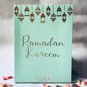 Pode incluir: Um calendário do Advento Ramadan Kareem verde menta com detalhes dourados. O calendário apresenta uma grelha de portas numeradas, com lanternas douradas decorativas no topo. As palavras "Ramadan Kareem" são escritas em ouro.