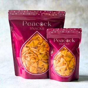 May include: Two bags of Peacock Snacks, one large and one small, are displayed. The bags are a deep magenta color with a white polka dot pattern at the top. The brand name is printed in white. The bags contain golden-colored, triangular snacks.