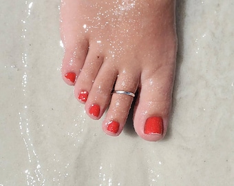 May include: A close-up of a foot with red nail polish and a silver toe ring. The foot is in shallow water on a white sandy beach.