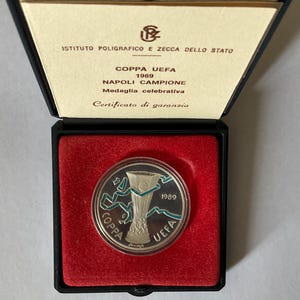 May include: A silver coin in a black box with a red velvet interior. The coin features a trophy design with the text "COPPA UEFA 1989" and "NAPOLI CAMPIONE". The box lid has text in Italian, including "ISTITUTO POLIGRAFICO E ZECCA DELLO STATO".