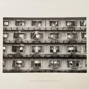 May include: Monochrome photograph of a building facade with many open windows. Some windows show people, others have washing hanging out. The image captures a scene of urban living.