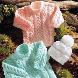 May include: A collection of hand-knitted baby clothing in soft pastel colors. The set includes a light pink sweater, a light blue cardigan with button closures, and a white knit hat with a pom-pom. The sweaters and hat feature a cable knit design.