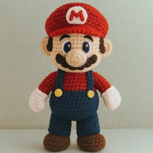 May include: A handmade crochet doll of the video game character Mario. The doll features a red hat with a white "M", a red shirt, blue overalls, and white gloves. The doll has a large mustache and is approximately 10 inches tall.