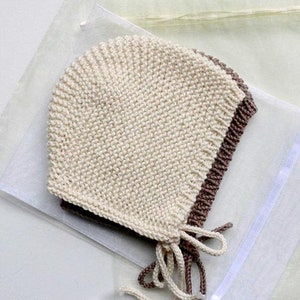 May include: A knitted bonnet in cream and brown. The bonnet is made of a soft, textured yarn and features a ribbed edge and ties. The bonnet is designed to keep a baby's head warm and protected.