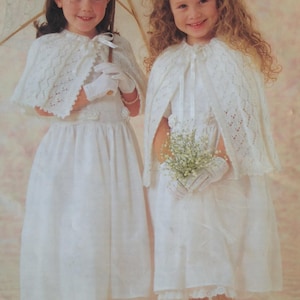 May include: Two young girls in white dresses and capes, holding a parasol and flowers. The dresses have lace details, and the girls wear white gloves and shoes. The background is a soft pink.