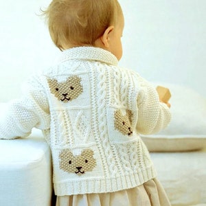 May include: A cream-coloured baby cardigan with a cable knit pattern and three embroidered bear faces on the back. The garment has a collared neckline and long sleeves. The baby is wearing a beige skirt.