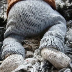 Könnte beinhalten: Nahaufnahme eines Babys in einem Strick-Outfit. Das Outfit umfasst graue Hosen und beige Babyschuhe. Das Oberteil des Outfits ist braun. Das Baby liegt auf einer flauschigen, grau-weißen Oberfläche.