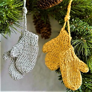 May include: Two knitted mitten decorations, one grey and one gold, suspended from a Christmas tree branch. The mittens are attached by yarn loops. Pine cones and evergreen needles create a festive background.