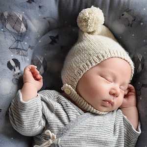 May include: A sleeping baby wearing a cream-colored knitted hat with a pom-pom and a gray striped onesie. The baby is lying on a gray patterned surface with hot air balloons.