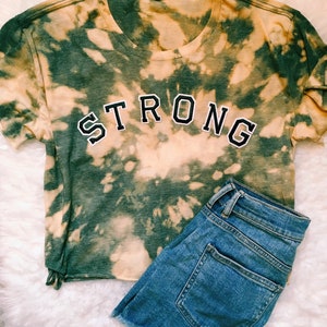 May include: A green and white tie-dye t-shirt with the word "STRONG" in black outlined letters. The shirt is paired with blue denim shorts.