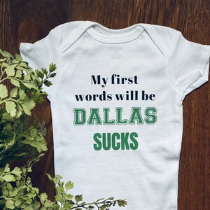 May include: White baby onesie with the text "My first words will be DALLAS SUCKS" printed in black and green.