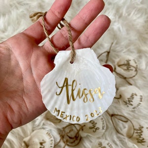 May include: White seashell ornament with a gold inscription that reads "Alissa Mexico 2023".