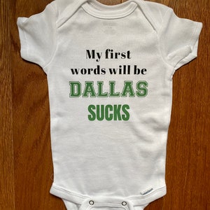 May include: White baby onesie with the text "My first words will be DALLAS SUCKS" in black and green lettering.