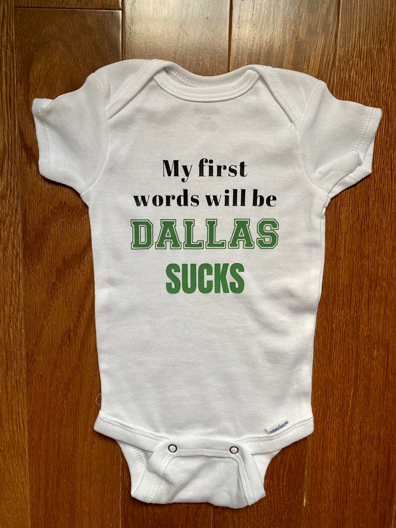 May include: White baby onesie with the text "My first words will be DALLAS SUCKS" in black and green lettering.