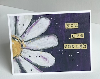 May include: A white daisy with a yellow centre on a purple background with gold glitter. The text "you are enough" is written in gold letters on the card.