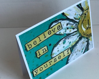 May include: A teal card with a white daisy painted on it. The card has the words "believe in yourself" written on it in yellow.
